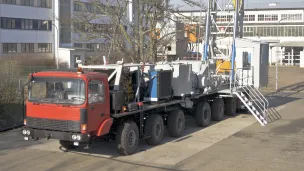 Large red and black mobile drilling truck parked near industrial buildings.