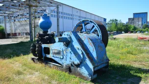 Rusty blue industrial machine on grass near a warehouse under a clear sky.