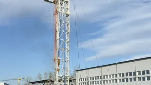 Tall drilling rig and yellow truck beside an industrial building under blue sky.