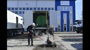 Two workers in safety gear use steaming equipment outside an industrial building.