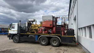 Large utility truck with machinery parked beside a white industrial building under cloudy sky.